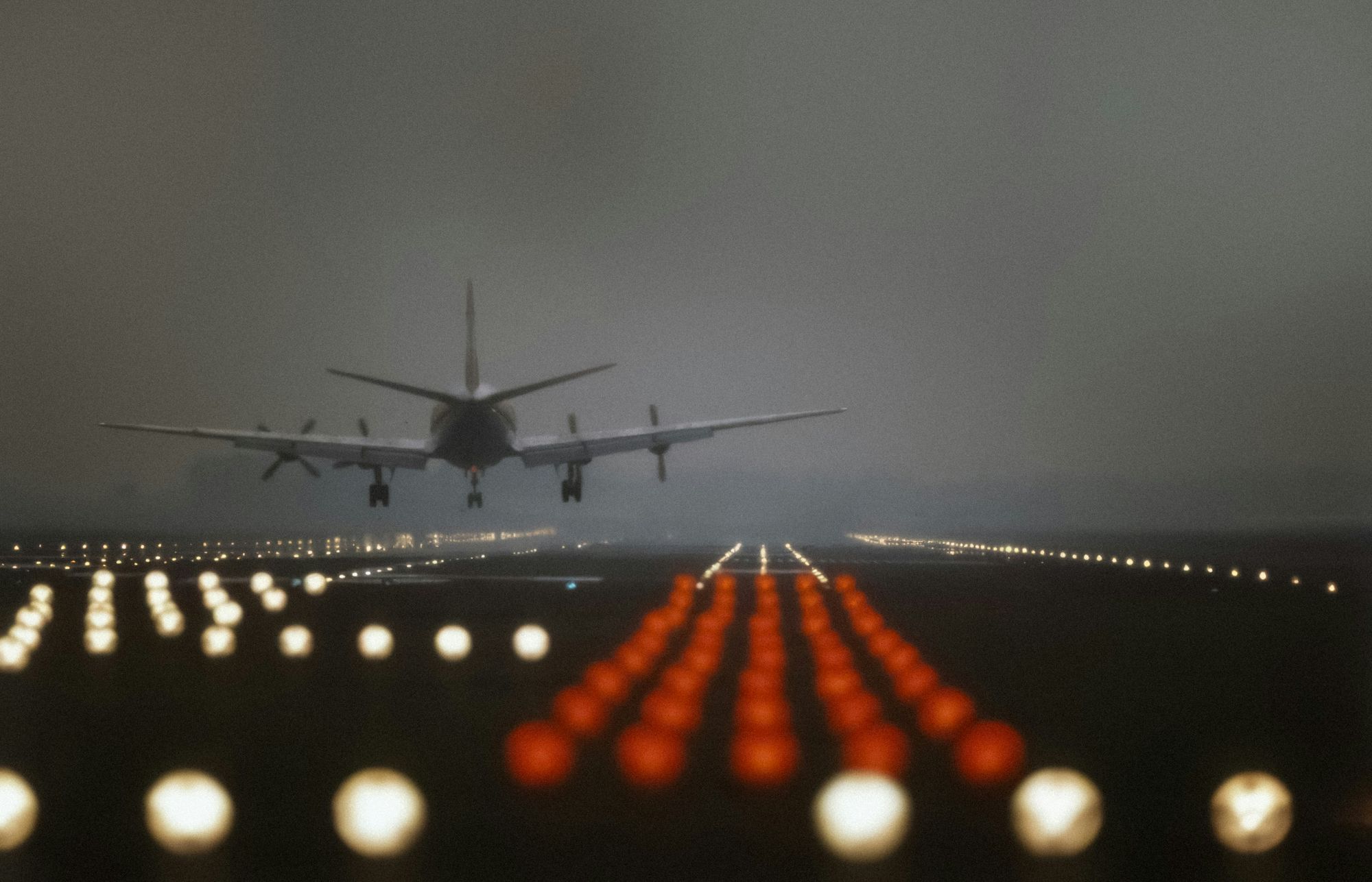 霧夜中一架客機放下起落架正要落地：跑道頭整排白色進場燈與紅色終端燈在暮色裡發光，機身化成剪影懸在燈海上方